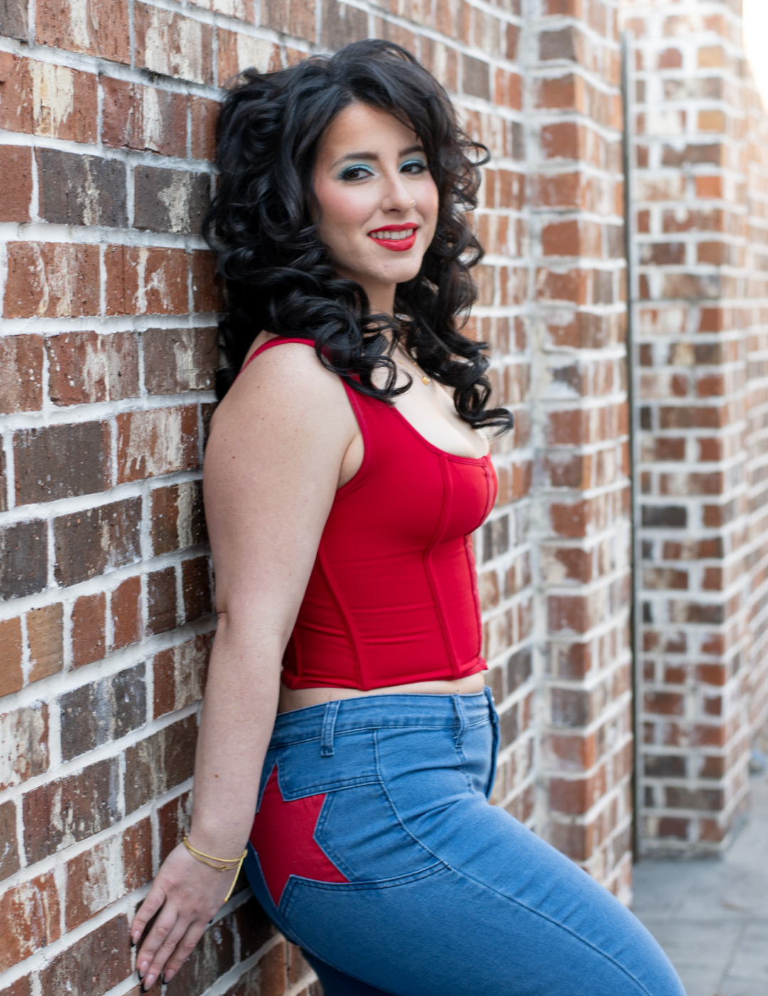 Hair stylist Emily West in Destrehan, Louisiana, with voluminous dark curls and a red top, posing against a brick wall.