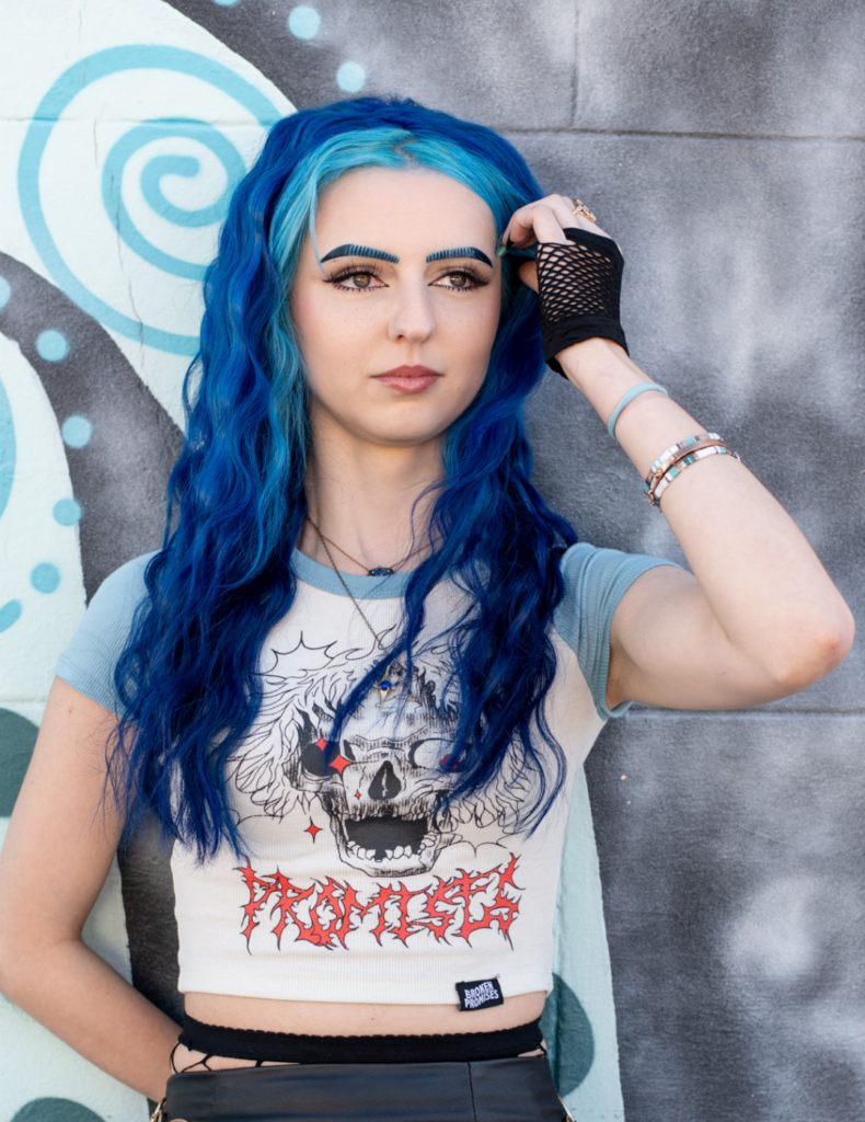 Nail artist Natalie Phillips in Destrehan, Louisiana, with vibrant blue hair and alt-style clothing, posing against a mural wall.