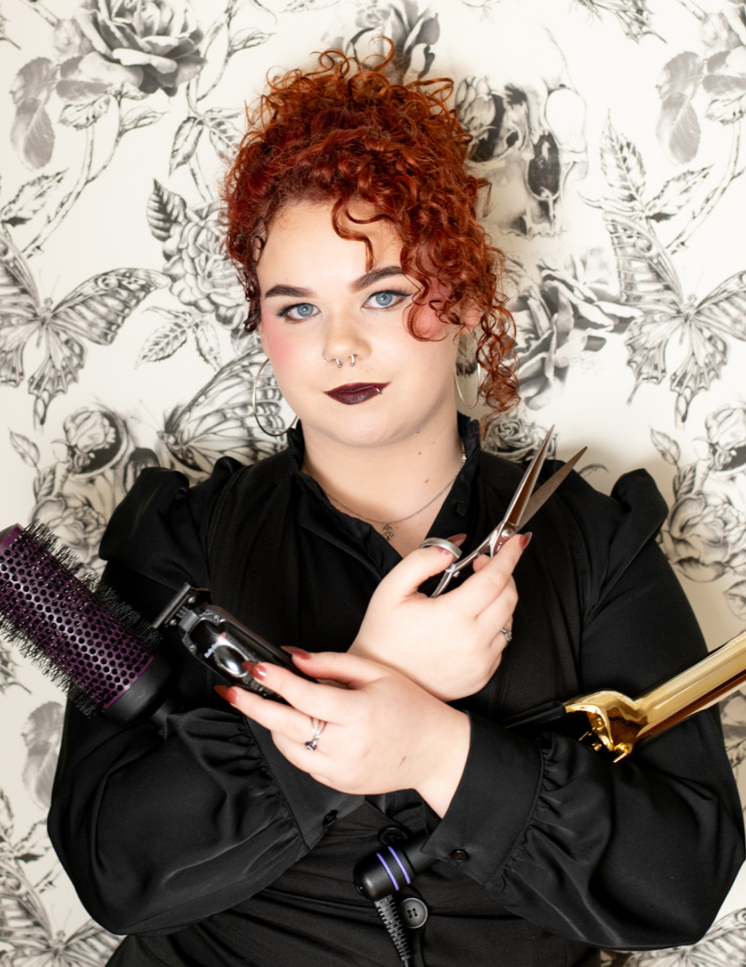 Hairstylist Shelby Gendusa in New Orleans, Louisiana, with curly red hair, holding professional styling tools including shears and a round brush.