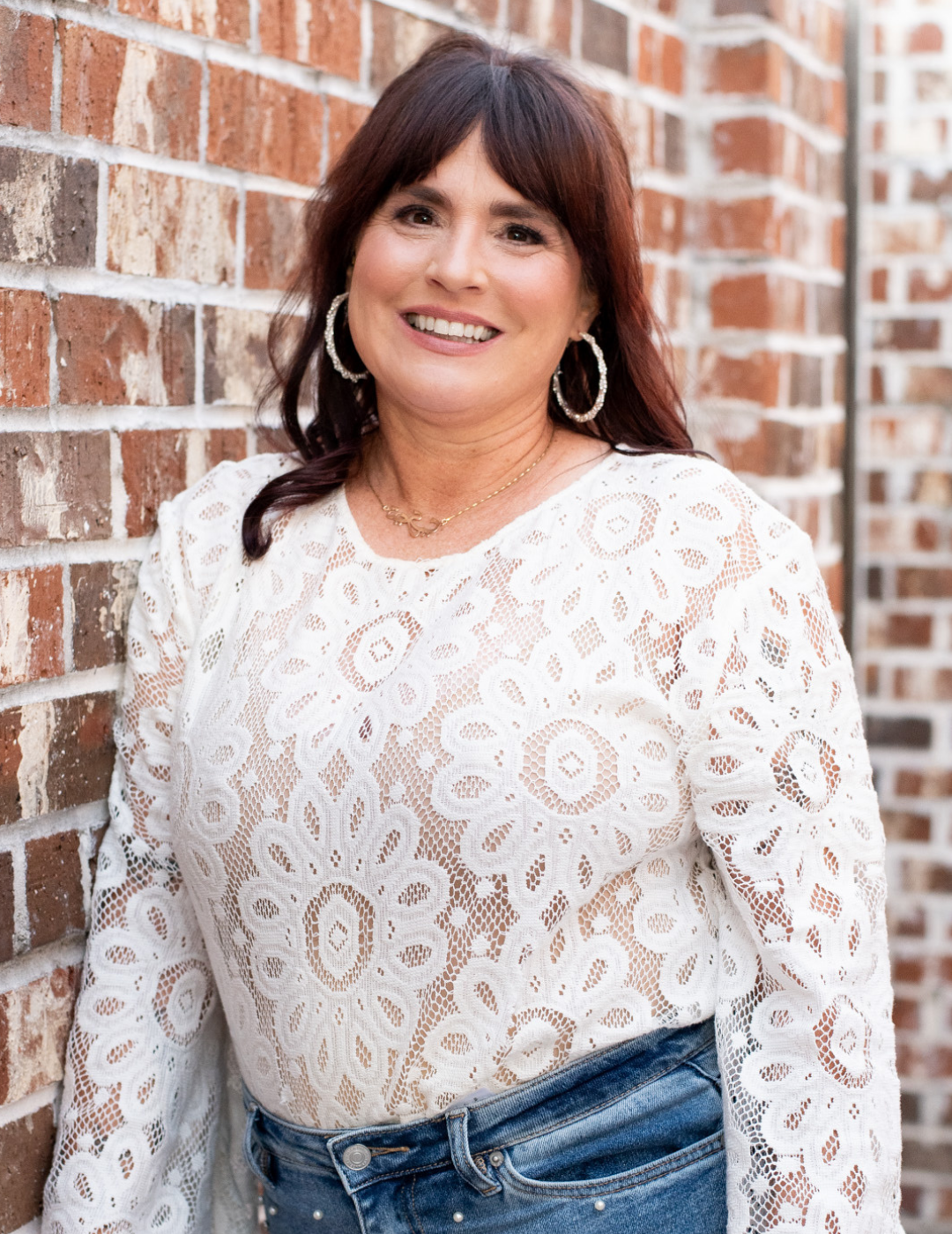 Salon owner Traci Woodard of TW Salon, a top-rated Louisiana salon, smiling in a white lace top against a brick background.