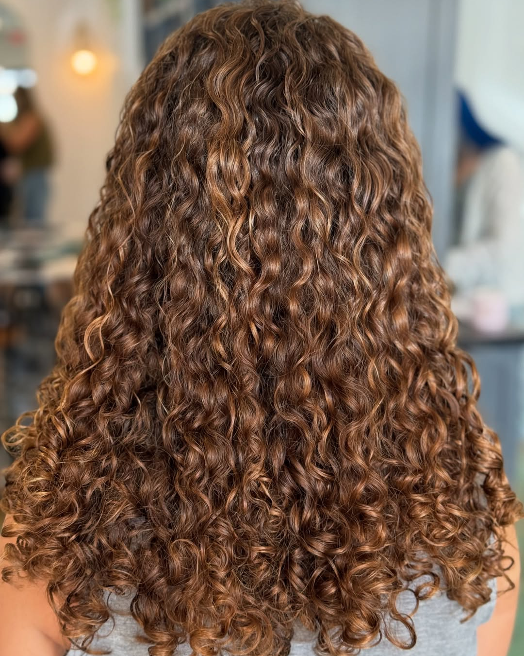 Back view of voluminous, tight brunette curls with golden brown highlights and a rounded shape, styled by Marie.