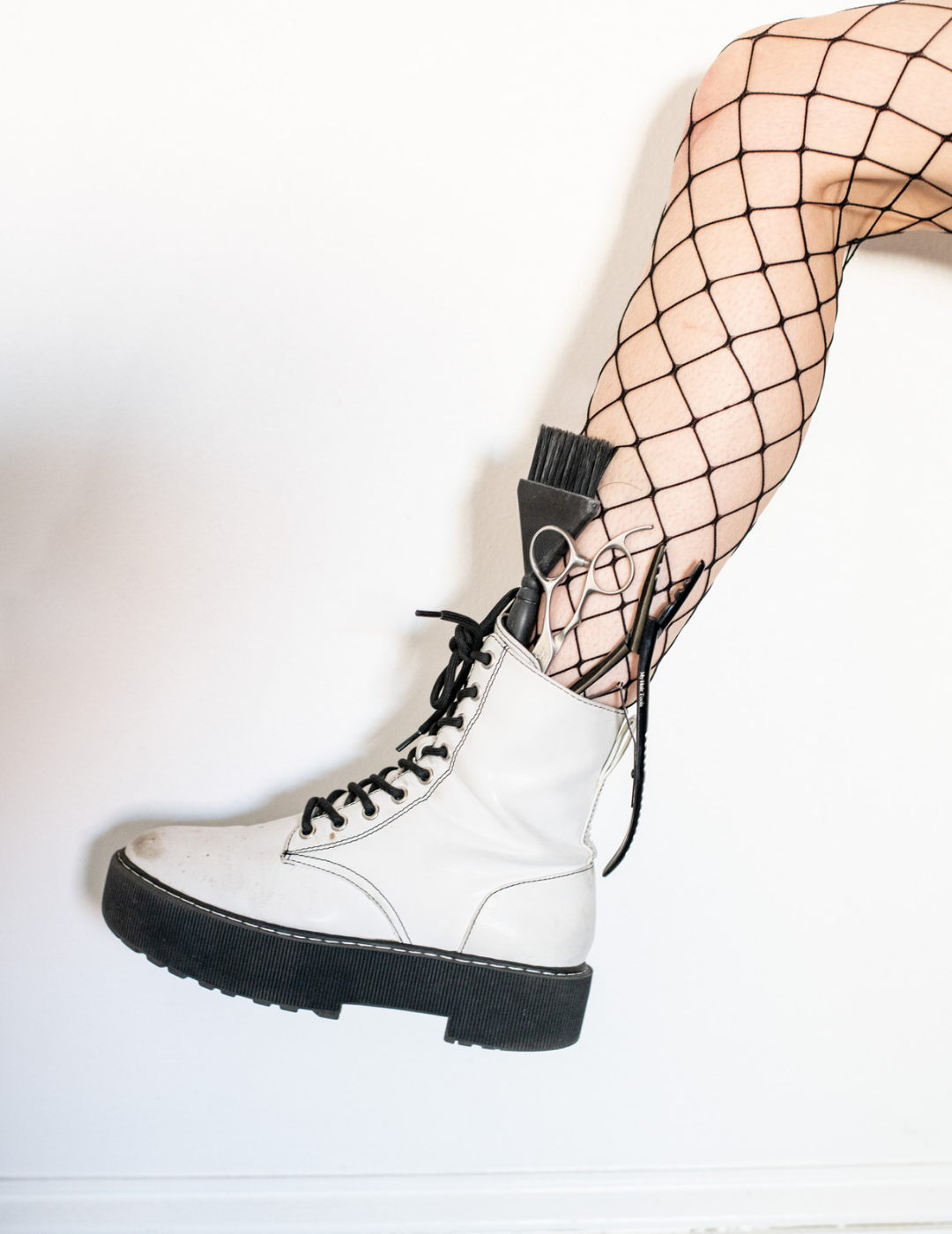 Close up of a model wearing fishnet stockings and white boots with hair shears and a color brush tucked into the boot.
