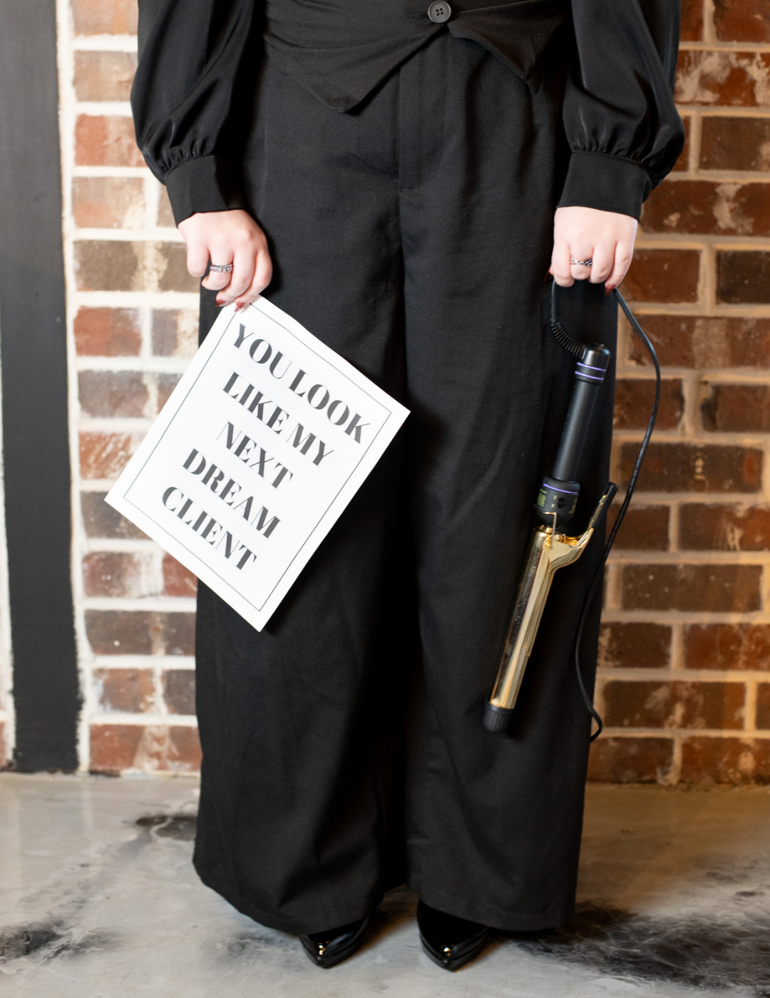 A stylist in professional black attire holding a curling iron and a sign that reads "You look like my next dream client in New Orleans, Louisiana.