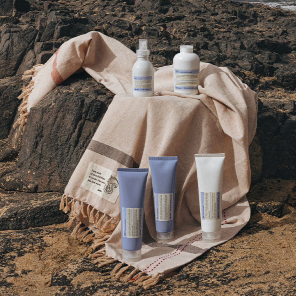 Davines SU hair care line for sun-exposed hair, featuring hair milk, body wash, and tan maximizer tubes on a beach towel over coastal rocks in New-Orleans, Louisiana.